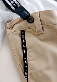 Beige trousers with a textured finish, featuring a black side label with white text, and navy suspenders attached at the waistband.