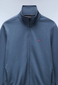 Donkerblauwe zip-up jas van stof, met een hoge kraag en een klein geborduurd vlaggetje op de borst. Gladde textuur en minimaal ontwerp.