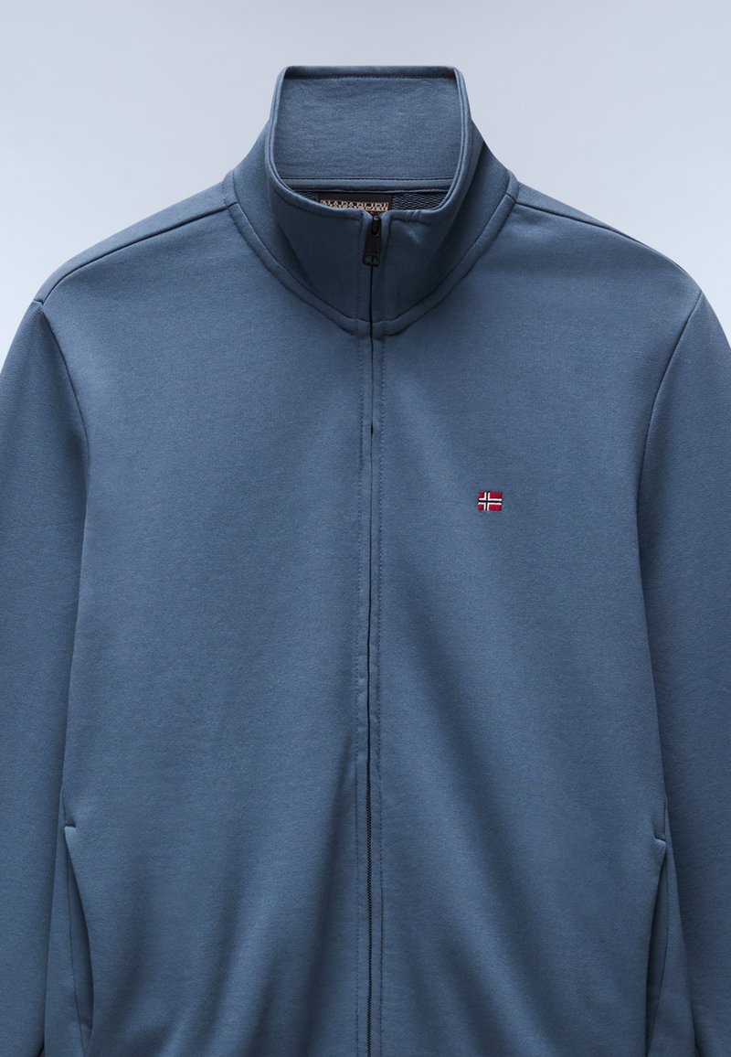 Chaqueta con cremallera en tela azul oscuro, con un cuello alto y un pequeño detalle de bandera bordado en el pecho. Textura suave y diseño minimalista.