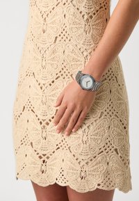 Montre en argent avec un cadran rectangulaire et des aiguilles des heures et des minutes noires, portée sur une main contre une robe crème à motif papillon en crochet.