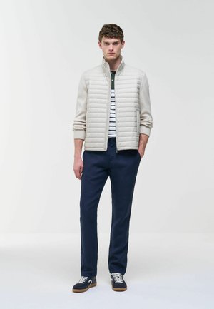 Fiatal férfi áll zsebre dugott kézzel, világos bézs színű steppelt dzsekit, csíkos inget, sötétkék nadrágot és fekete, fehér fűzőjű tornacipőt viselve.