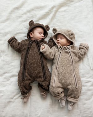 Two babies in cozy, bear-themed onesies: one dark brown and the other light beige, both with ears and contrasting zippers. Soft fleece texture.