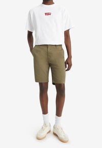 T-shirt blanche à manches courtes avec un logo rouge Levi's, associée à un short vert olive. Les chaussures sont des baskets beige clair avec des semelles blanches et des chaussettes.