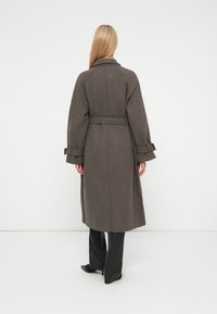 Blonda sieviete stāv, ģērbusies garā brūnā apmetnī ar jostu un piesietiem aprocēm, melnās biksēs un melnās papēžu kurpēs, vērsta pret baltu sienu.