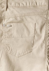 Beige bakficka på byxor med synlig sömnad, bälteshällor, metallnitar och liten rektangulär varumärkesetikett.