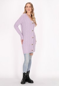 Lavendel kabelgebreide cardigan met vijf gouden knopen, v-hals, lange mouwen en een knielange snit, gedragen over versleten lichtblauwe jeans.
