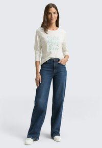 Vrouw in een witte blouse met lange mouwen en abstracte print, een donkerblauwe wijde jeans en witte sneakers, staand met één hand in de zak.