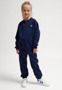 Mörkblå sweatshirt och matchande joggers i mjukt material, med en liten broderad logotyp på bröstet, ihop med vita sneakers.