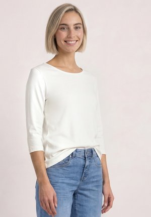 Ung kvinde med kort blondt hår iført en hvid langærmet top og blå jeans, smilende, stående mod en ensfarvet lys baggrund.