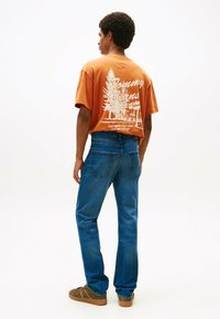 Hombre de pie de lado, vestido con una camiseta gráfica naranja que dice "Tommy Jeans" y unos vaqueros azules, con zapatillas marrones, sobre un fondo blanco.