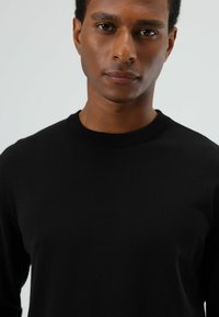 Zwarte trui met lange mouwen en ronde hals, gemaakt van zachte stof. Voorzien van geribbelde halslijn en aansluitende pasvorm. Gladde textuur met minimale details.