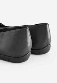 Musta nahkainen tasapohjainen kenkä, jossa on sileä pinta, pyöreä korko ja hieman kohotettu pohja. Suunnittelussa on puhdas, minimalistinen ilme.