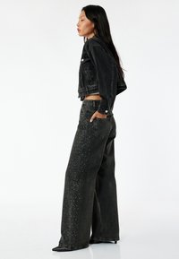 Sort denimjakke med cropped design og lange ærmer, parret med højtaljede bukser med brede ben, som har rhinsten-accenter langs det ene ben.