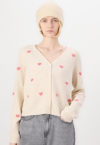Cárdigan de punto en beige con patrones de corazones en rosa claro, escote en V, cierre frontal con botones y puños acanalados. Combinado con un gorro beige a juego.