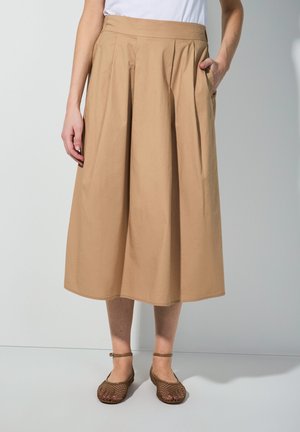 Beige Plissée-Rock mit breitem Bund und Seitentaschen, aus glattem Stoff gefertigt. Kombiniert mit braunen, offenen Wedge-Sandalen.