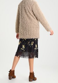 Personne portant une veste longue à manches longues en peluche beige, une jupe midi noire à motifs floraux et des bottines brunes, se tenant devant un fond uni.