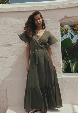 Junge Frau mit langem Haar, die ein olivgrünes Wickelkleid mit kurzen Ärmeln und eine Muschelkette trägt, steht vor einer weißen, strukturierten Wand.