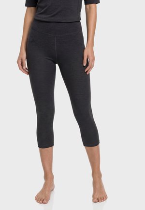 Frau trägt schwarze kurze Leggings und ein schwarzes Oberteil, steht barfuß vor einem schlichten hellen Hintergrund.