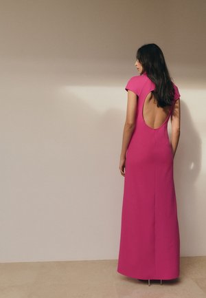 Mujer con cabello largo y oscuro, vestida con un vestido rosa brillante hasta el suelo con una gran abertura en la espalda, de pie contra una pared beige lisa.