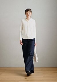 Weißes langärmliges Button-Up-Hemd, dunkelblaue weite Jeans und schwarze Schuhe. Das Model trägt eine kleine weiße Handtasche. Einfache, klare Linien.