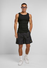 Svart tanktop, svarte shorts og hvite joggesko. Shortsene har en lomme og logo. Personen har briller og en slank kroppsbygning.