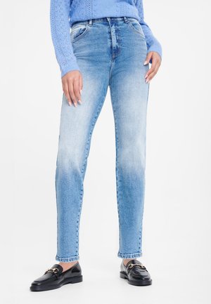 Person trägt hellblaue High-Waist-Jeans, einen blauen Strickpullover und schwarze Leder-Loafers und steht vor einem weißen Hintergrund.