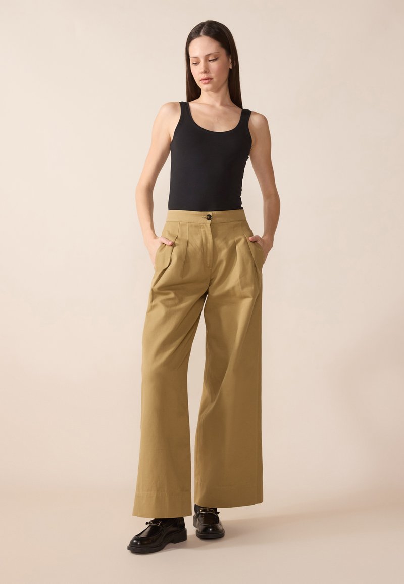 Široké khaki kalhoty s sklady a bočními kapsami, doplněné o těsně přiléhající černý top na ramínka. Modelka stojí s rukama v kapsách.