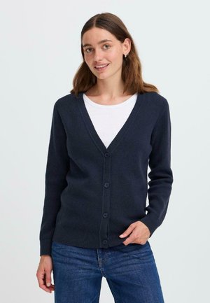 Cardigan navy en matériau doux avec un col en V, six boutons sur le devant et des poignets côtelés, accompagné d'une chemise blanche en dessous.