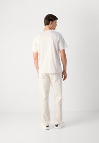 Mand iført en ensfarvet lys beige t-shirt, matchende bukser og hvide sneakers, står med ryggen til kameraet foran en hvid baggrund.