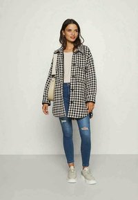 Melnbalts houndstooth rakstainais lielgabarīta mētelis, pogājamā stila ar sānu kabatām, apvienots ar bēšiem topiem, zilām skinny džinsām un pelēkiem sporta apaviem.