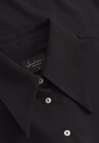 Camicia nera con colletto appuntito, placca con bottoni in argento e una texture liscia. L'etichetta riporta "Vascha, Stoccolma, S."