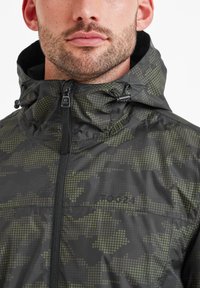Black and green camouflage patterned jacket with a hood, front zipper, drawstrings, and mesh-lined collar. Features a brand logo on the chest.