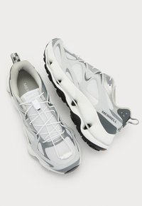 Adidași albi și gri cu partea superioară din plasă, suprapuneri texturate și talpă neagră. Prezintă șireturi elastice și o bandă de prindere pe călcâi.