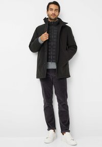 Sort vandtæt jakke med knap- og lynlåslukning, parret med en sort dunvest, grå sweater og mørke fløjlsbukser. Hvide sneakers.