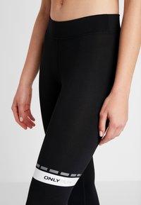 Närbild av person som bär svarta tights med hög midja och ett vitt band märkt "ONLY PLAY" nära låret.