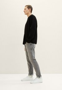 Suéter negro de manga larga, jeans grises y zapatillas blancas. Diseño simple con un corte relajado y textura lisa. Perfil lateral sobre un fondo claro.