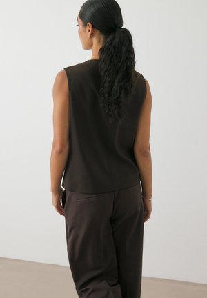 Brown, sleeveless top with a high neckline, made of smooth fabric, paired with loose brown trousers; textured hair tied back.