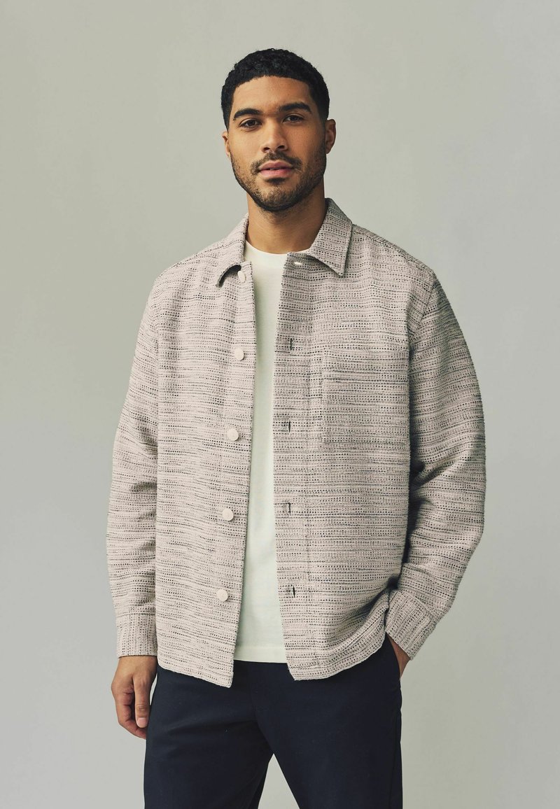 Light gray woven jacket with a textured pattern, front pockets, and light buttons, worn over a cream shirt and dark pants.