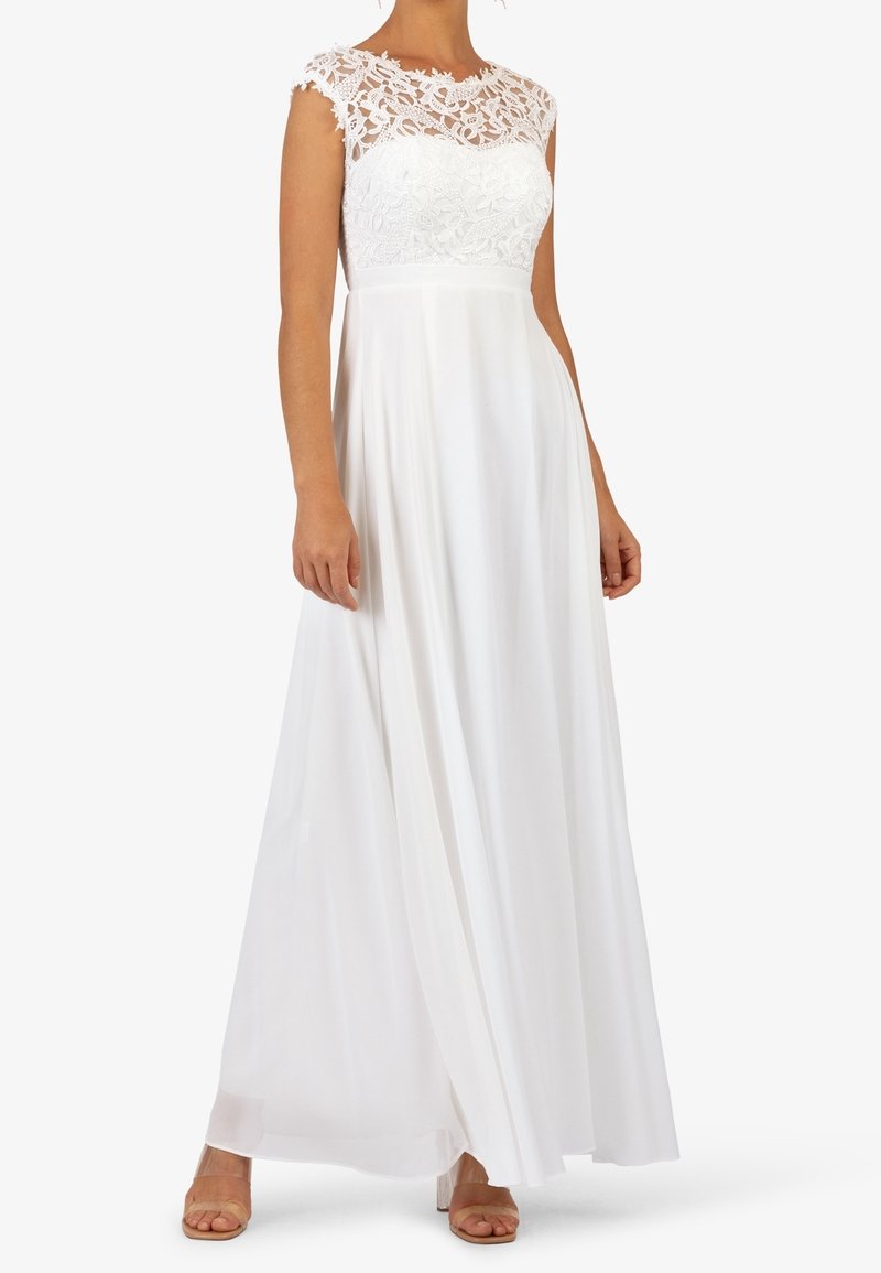Femme portant une robe blanche longue sans manches avec un corsage en dentelle florale et un décolleté en dentelle transparente, associée à des talons à brides transparentes.