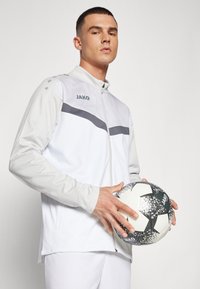 Jachetă sport albă cu accente gri, front cu fermoar, țesătură texturată și logo. Ținând o minge de fotbal cu un model alb-negru.