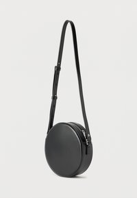 Μαύρη στρογγυλή τσάντα μέσω ώμου με ρυθμιζόμενο λουρί και επάνω κλείσιμο με φερμουάρ, εκτεθειμένη σε απλό λευκό φόντο.