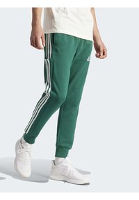 Gröna sweatpants med tre vita sidostripes, elastiska muddar och en Adidas-logga. Bärs med vita sneakers och en ljus färgad topp.