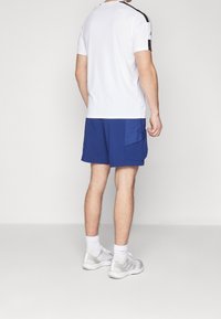 Outfit atletico da uomo composto da una maglietta bianca a maniche corte, pantaloni corti blu con tasca laterale e scarpe sportive grigie con calze bianche.