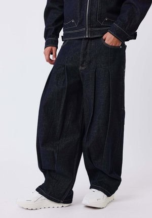 Personne portant un pantalon en jean large et foncé ainsi qu'une veste assortie, debout avec une main dans la poche, baskets blanches visibles sous le pantalon.