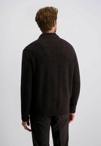 Man die met zijn rug naar de camera staat, gekleed in een donkerbruin sweater en zwarte broek, tegen een effen lichte achtergrond.