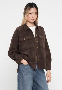 Braunes Cordhemd mit zwei Brusttaschen, Knopfverschluss und spitzem Kragen, kombiniert mit hellblauen Jeans.