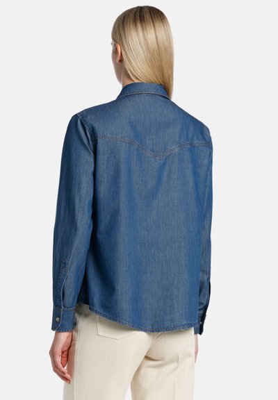 Denim srajca v temno modri barvi, z izpostavljenim ovratnikom in gumbi na manšetah. Hrbtna stran ima yoke dizajn s subtilno V-oblikovano šivno podrobnostjo.