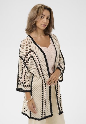 Ženska z ramensko dolžino las, oblečena v bež kvačkano jopico s črnim robom čez rožnato čipkasto majico in kremasto krilo, pozira pred belim ozadjem.