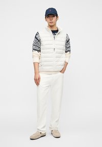 Φουσκωτό λευκό γιλέκο πάνω από μπλε πουά πουλόβερ, κρεμ παντελόνι και μπεζ μοκασίνια. Μπλε καπέλο με λογότυπο ολοκληρώνει την εμφάνιση. Απλά στοιχεία σχεδίασης.