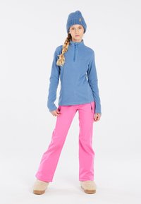 Lichtblauwe fleece pullover met ritskraag, gecombineerd met felroze skibroeken met ritssluitingen en beige fluffy slippers.